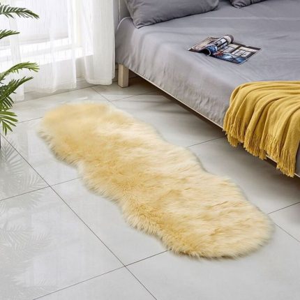 Tapis long en fausse fourrure beige de forme irrégulière posé sur un sol carrelé blanc à côté d'un lit.