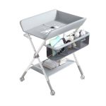 Table à langer pliable pour bébé montée sur des pieds en métal en X avec des roulettes, équipée d'un matelas à langer avec ceinture de sécurité, d'un panier de rangement latéral compartimenté et d'une étagère inférieure en tissu.
