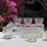 Ensemble de plusieurs verres transparents aux parois finement striées et de forme évasée, dotés d'une bague brillante texturée à la base, disposés sur une table blanche devant un arrière-plan floral.