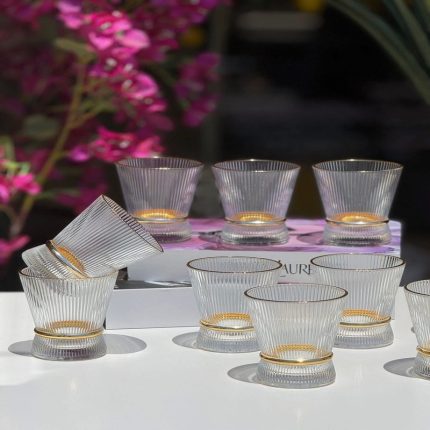 Ensemble de plusieurs verres transparents aux parois finement striées et de forme évasée, dotés d'une bague brillante texturée à la base, disposés sur une table blanche devant un arrière-plan floral.