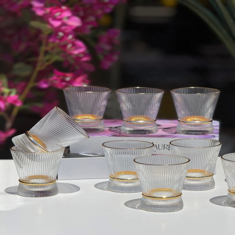 service-verres-eau-stries-canneles Ensemble de plusieurs verres transparents aux parois finement striées et de forme évasée, dotés d'une bague brillante texturée à la base, disposés sur une table blanche devant un arrière-plan floral.