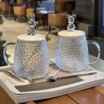 Deux mugs en verre texturé martelé avec des couvercles surmontés de figurines de lapin et d'ours de style métallique, accompagnés de cuillères assorties.