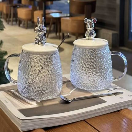 Deux mugs en verre texturé martelé avec des couvercles surmontés de figurines de lapin et d'ours de style métallique, accompagnés de cuillères assorties.