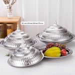 Trois plats de service ronds en acier inoxydable brillant, empilés et surmontés de couvercles bombés et ciselés avec poignées décoratives, accompagnés d'un plat ouvert présentant des fruits frais comme des caramboles et des raisins.