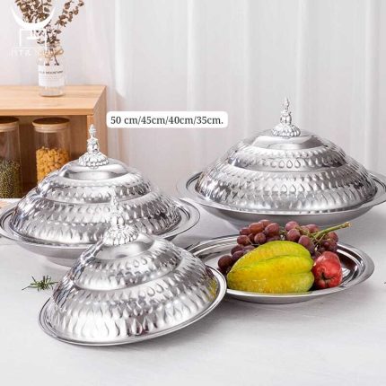 Trois plats de service ronds en acier inoxydable brillant, empilés et surmontés de couvercles bombés et ciselés avec poignées décoratives, accompagnés d'un plat ouvert présentant des fruits frais comme des caramboles et des raisins.