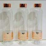 Trois bouteilles cylindriques en verre transparent alignées, décorées de trois bandes horizontales or rose à leur base et équipées de bouchons métalliques assortis.