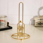 Support vertical porte-essuie-tout en métal doré brillant, doté d'une tige centrale haute et d'une arche frontale décorée de deux fleurs ciselées, posé sur un plan de travail de cuisine blanc.
