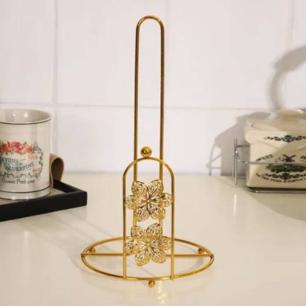 Support vertical porte-essuie-tout en métal doré brillant, doté d'une tige centrale haute et d'une arche frontale décorée de deux fleurs ciselées, posé sur un plan de travail de cuisine blanc.