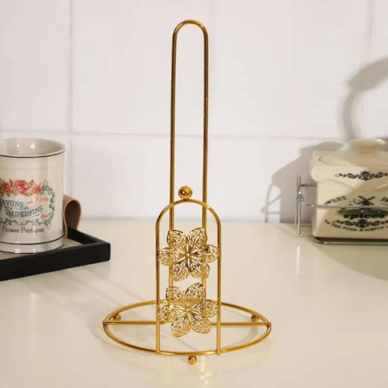 porte-essuie-tout-metal-dore-decoratif-fleurs Support vertical porte-essuie-tout en métal doré brillant, doté d'une tige centrale haute et d'une arche frontale décorée de deux fleurs ciselées, posé sur un plan de travail de cuisine blanc.