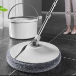 Balai de nettoyage avec une tête rotative circulaire blanche dotée de franges en microfibre grise, posé sur un sol carrelé foncé près de son seau d'essorage.