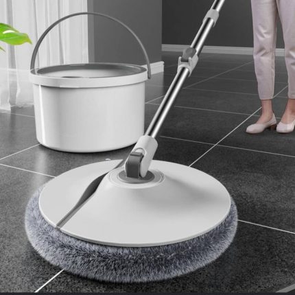 Balai de nettoyage avec une tête rotative circulaire blanche dotée de franges en microfibre grise, posé sur un sol carrelé foncé près de son seau d'essorage.