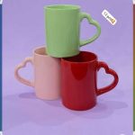 Trois mugs colorés vert, rose et rouge avec une anse originale en forme de cœur sur un fond violet.